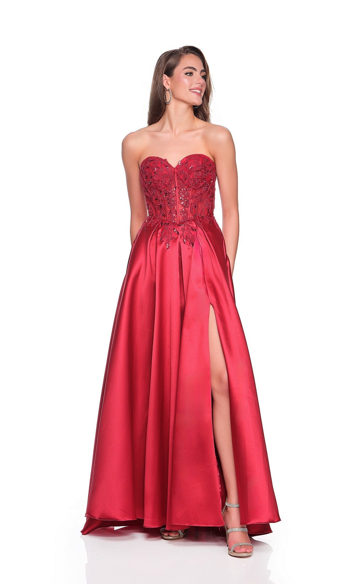 A woman stands against a plain white background wearing the Long Prom Dress: Dave and Johnny 12575, a strapless red gown with a corset bodice, floral embellishments, floor-length satin skirt with high slit, and silver heels.